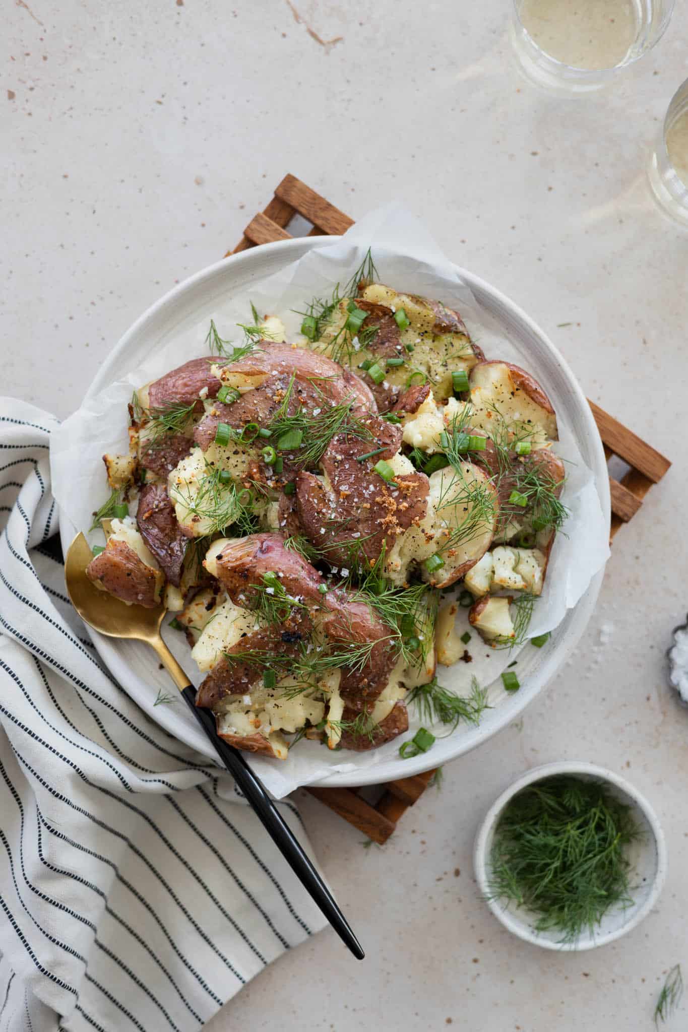 Smashed potatoes with green onions and dill on a platter with serving utensils and a striped linen. 