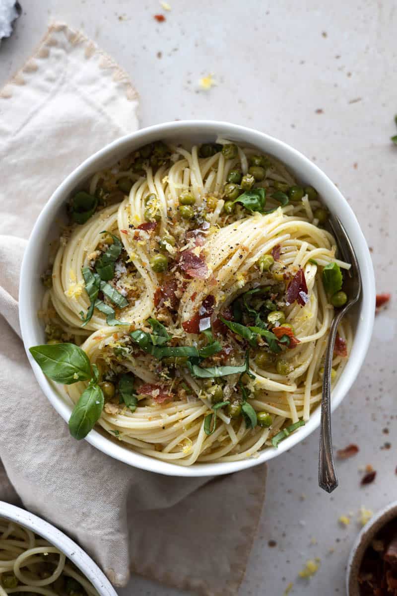 A bowl of spring pasta with lemon, peas and prosciutto with a spoon and a cream colored linen. 