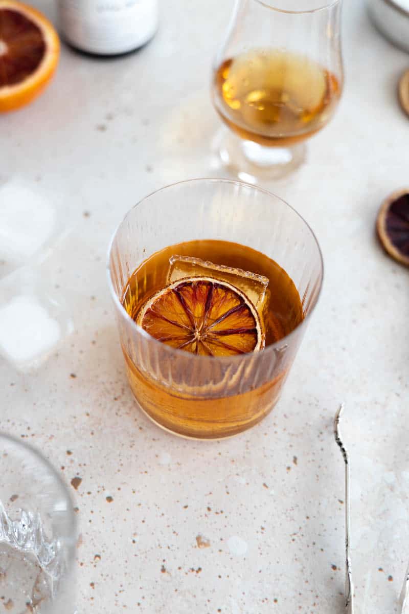 A Blood Orange Old Fashioned in a cocktail glass with a large ice cube and one dried blood orange slice. 