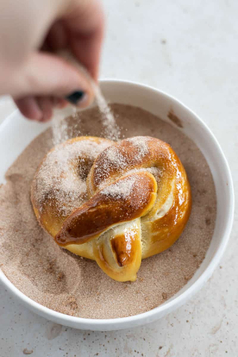 Sprinkling a baked pretzel in a bowl of cinnamon sugar.