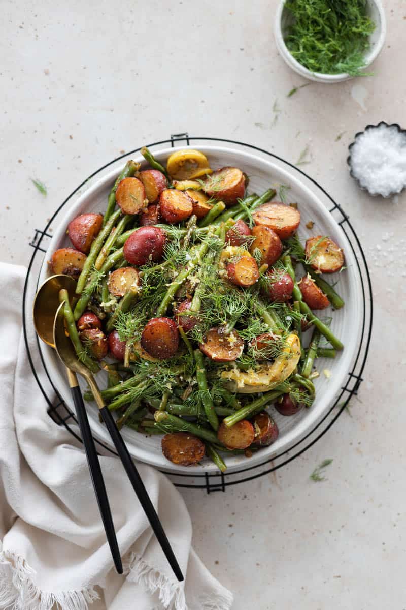 A serving platter of Green Beans and Potoates on a table with serving utensils. 