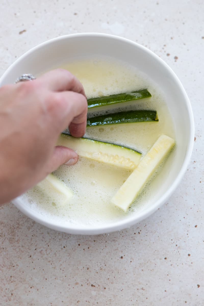 A hand tossing zucchini slices in a bowl of egg whites. 