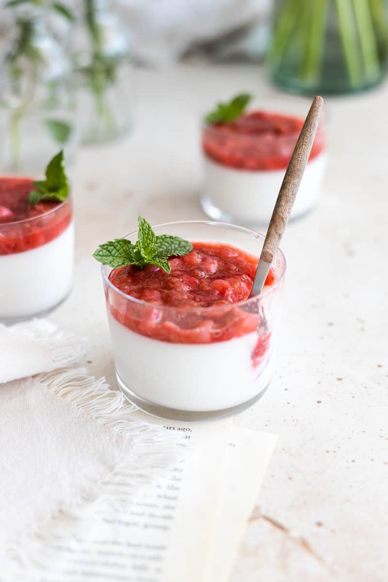 Panna cotta in a glass cup with strawberry rhubarb compote on top and a mint sprig garnish. A spoon is set in the glass to take a scoop. 