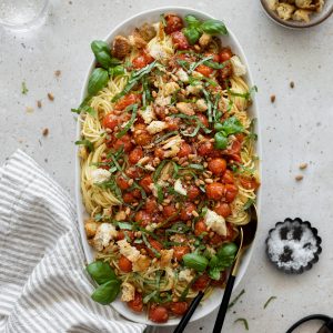 Pasta with burst cherry tomatoes on a serving platter, topped with basil, bread chunks and pine nuts.