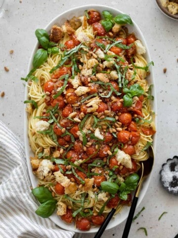 Pasta with burst cherry tomatoes on a serving platter, topped with basil, bread chunks and pine nuts.