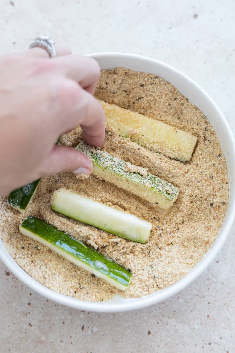A hand tossing zucchini slices in a bowl with the breadcrumb mixture. 