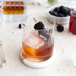 A Blackberry Old Fashioned in a cocktail glass with a large ice cube and blackberries on a cocktail pick.