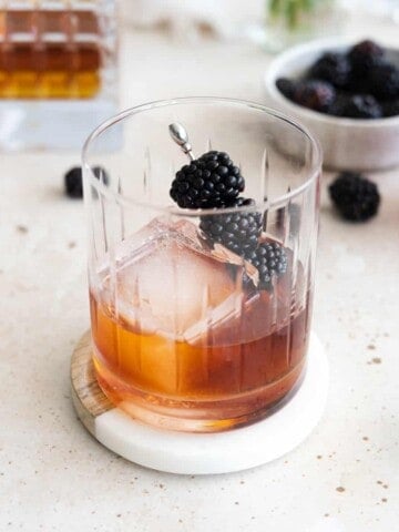 A Blackberry Old Fashioned in a cocktail glass with a large ice cube and blackberries on a cocktail pick.