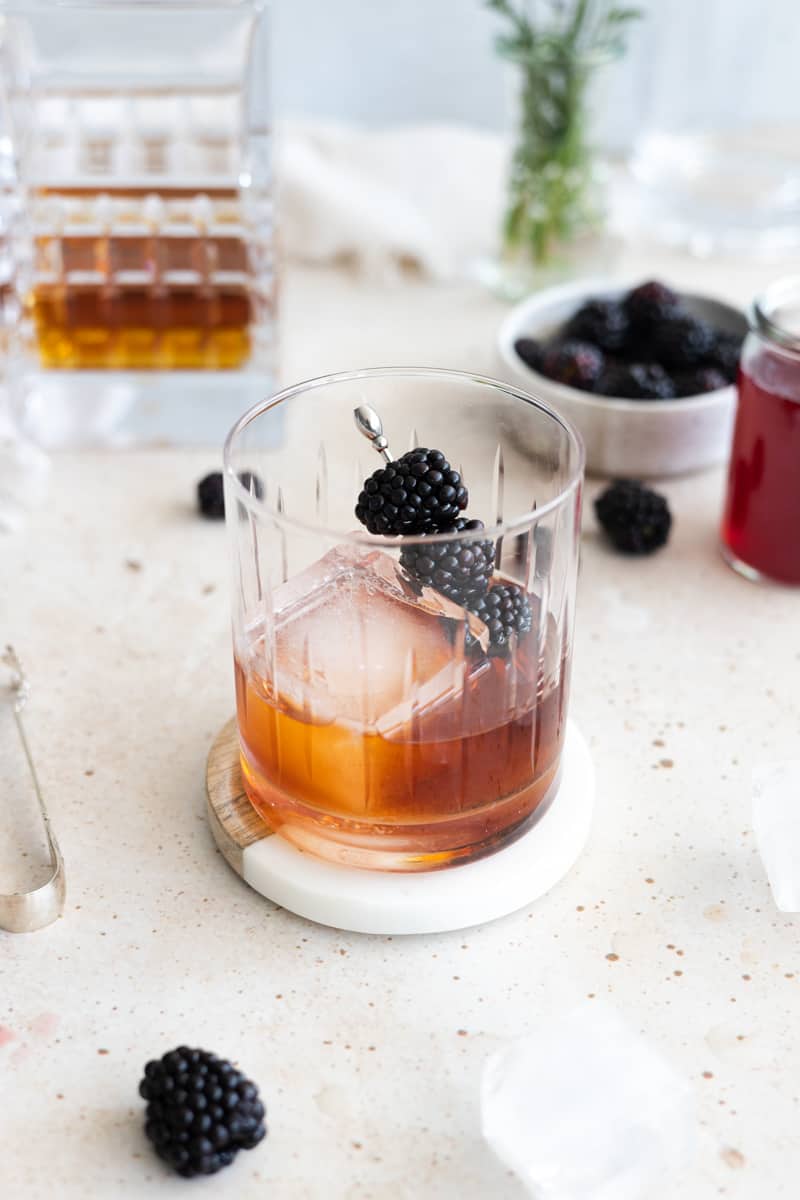 A Blackberry Old Fashioned in a glass with a large ice cube and blackberries on a cocktail pick.