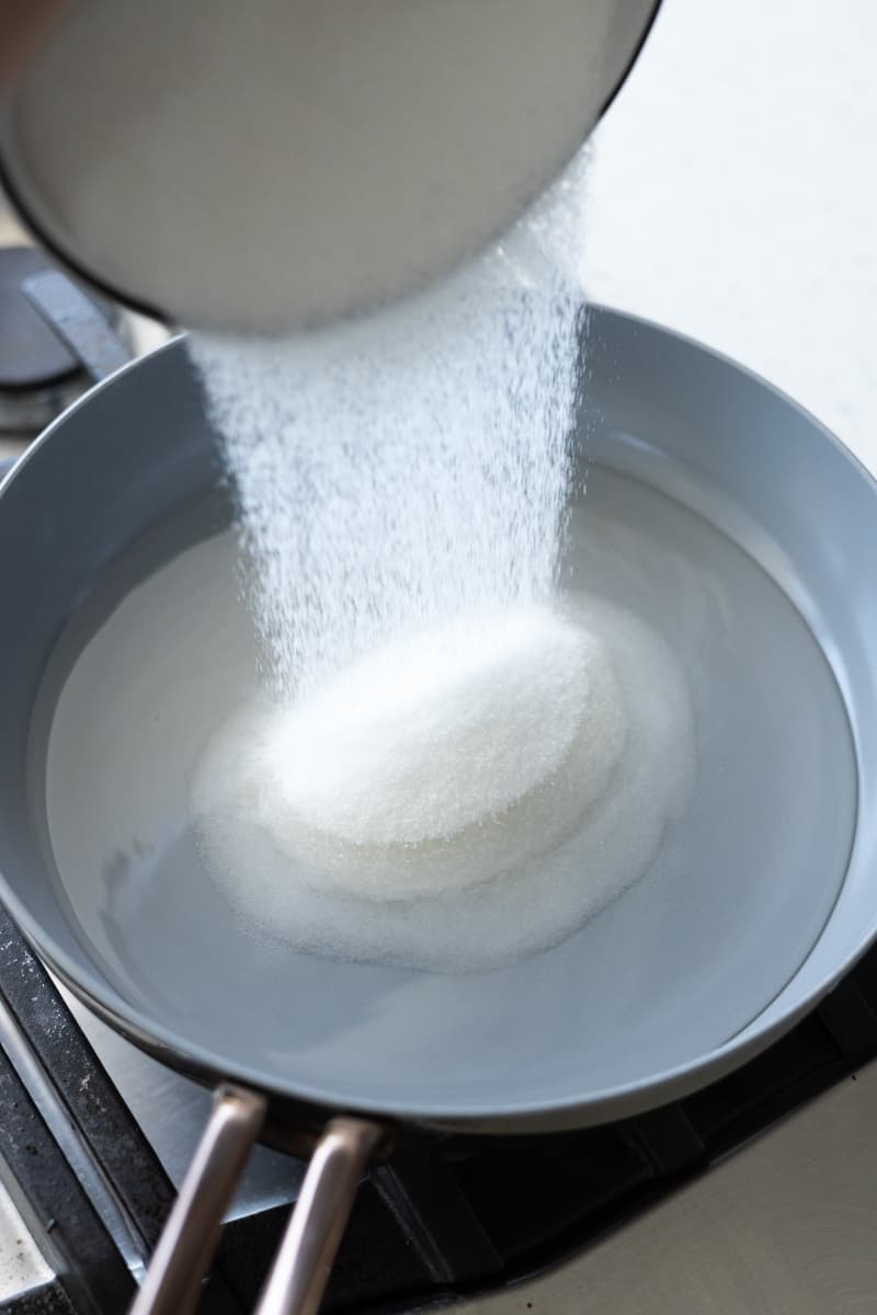 Granulated sugar being poured into the water in a small skillet. 