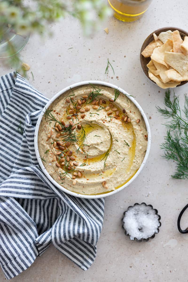 A bowl of lemon dill hummus with a small bowl of pita chips and a blue and white linen. 