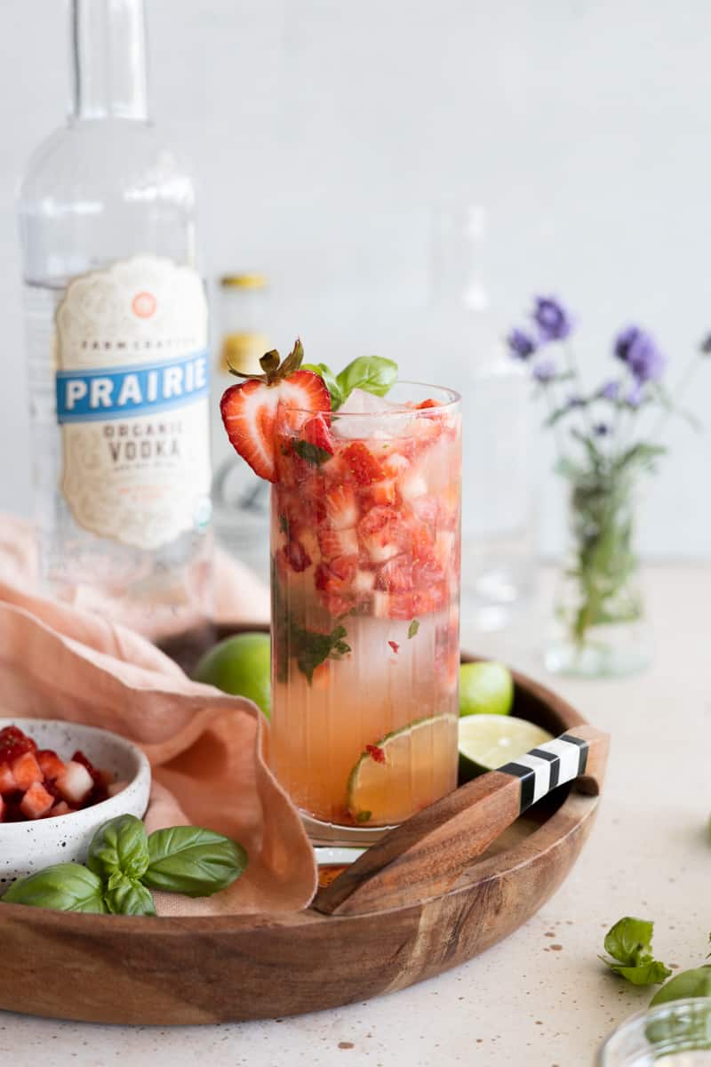 A strawberry basil vodka cocktail garnished with a strawberry slice on a wooden tray. 