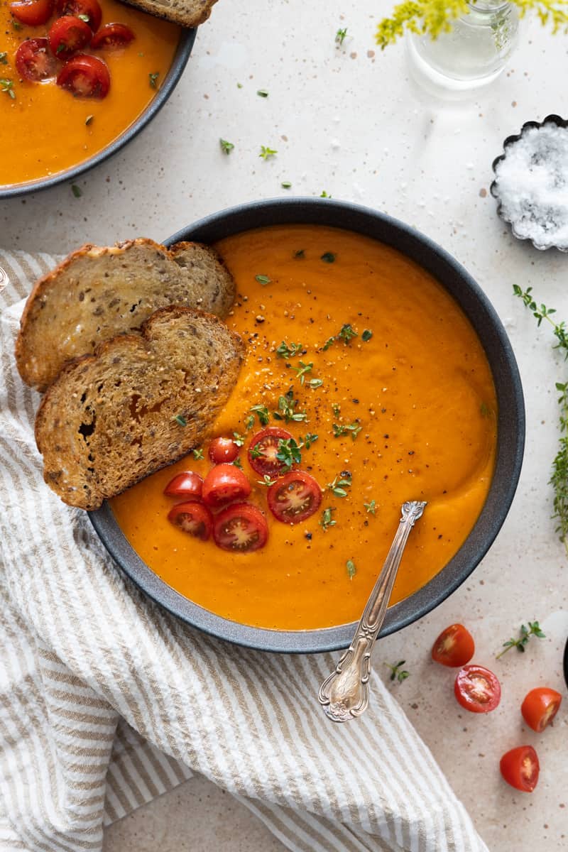 Butternut squash and red pepper soup in a grey bowl with two slices of crispy bread, halved cherry tomatoes and fresh thyme. 