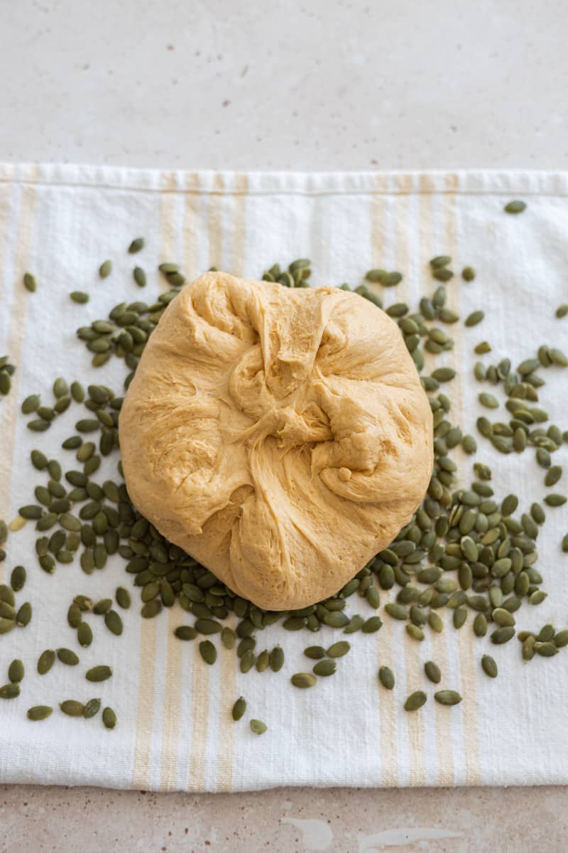 Dough ball on top of a kitchen towel spread with pepita seeds. 