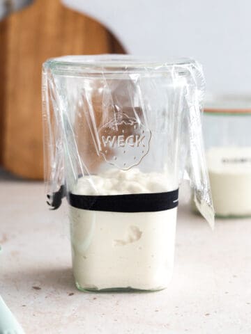 Sourdough starter in a tall glass jar covered with plastic wrap.