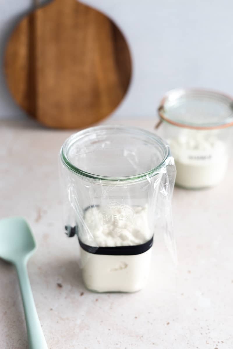 Sourdough starter in a tall glass jar with a black rubberband, covered in plastic wrap. 