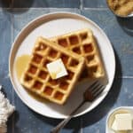 Cropped image of two Sourdough Discard Waffles on a white plate with a pad of butter and maple syrup.