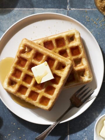 Cropped image of two Sourdough Discard Waffles on a white plate with a pad of butter and maple syrup.