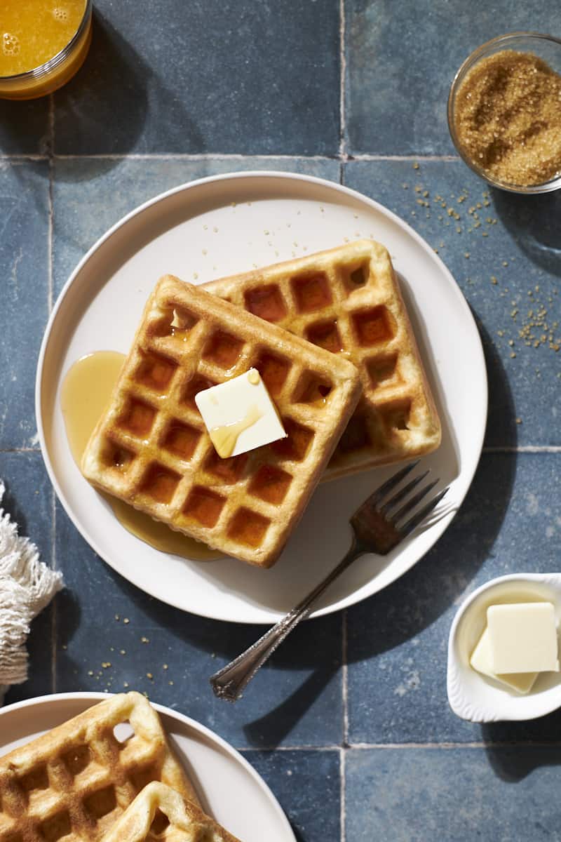 Overhead view of two Sourdough Discard Waffles on a white plate with a pad of butter and maple syrup.