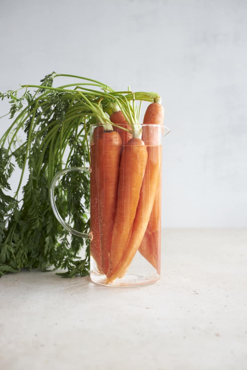 Carrots with green stems submerged in a glass pitcher of water.