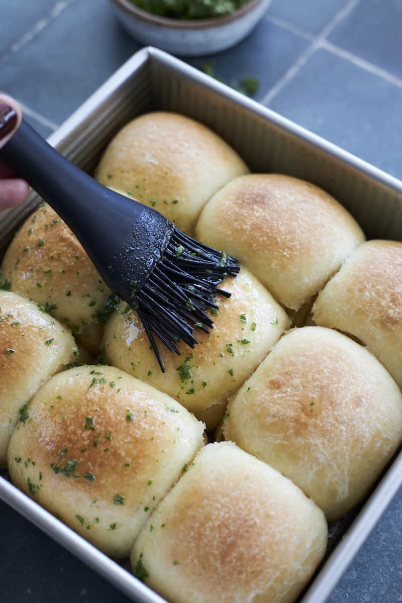 A brush putting melted butter and fresh parsley on garlic rolls.