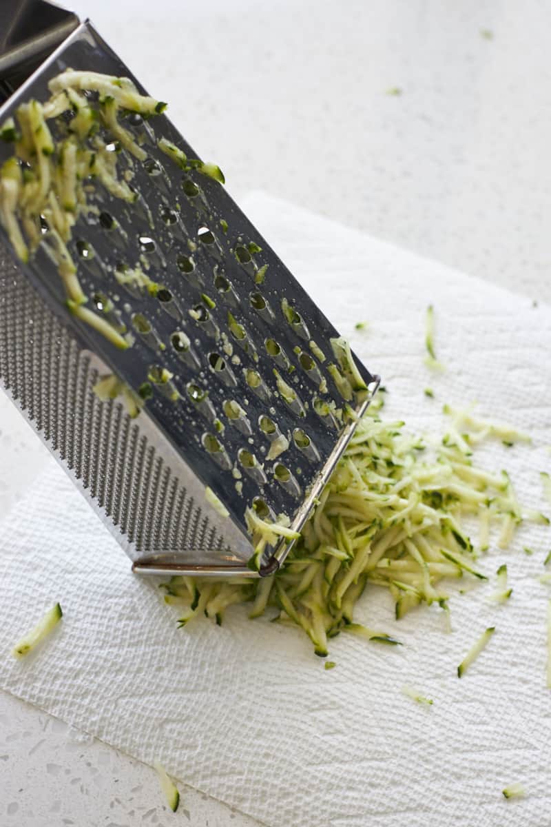 Zucchini being shredded on a paper towel with a box grater. 
