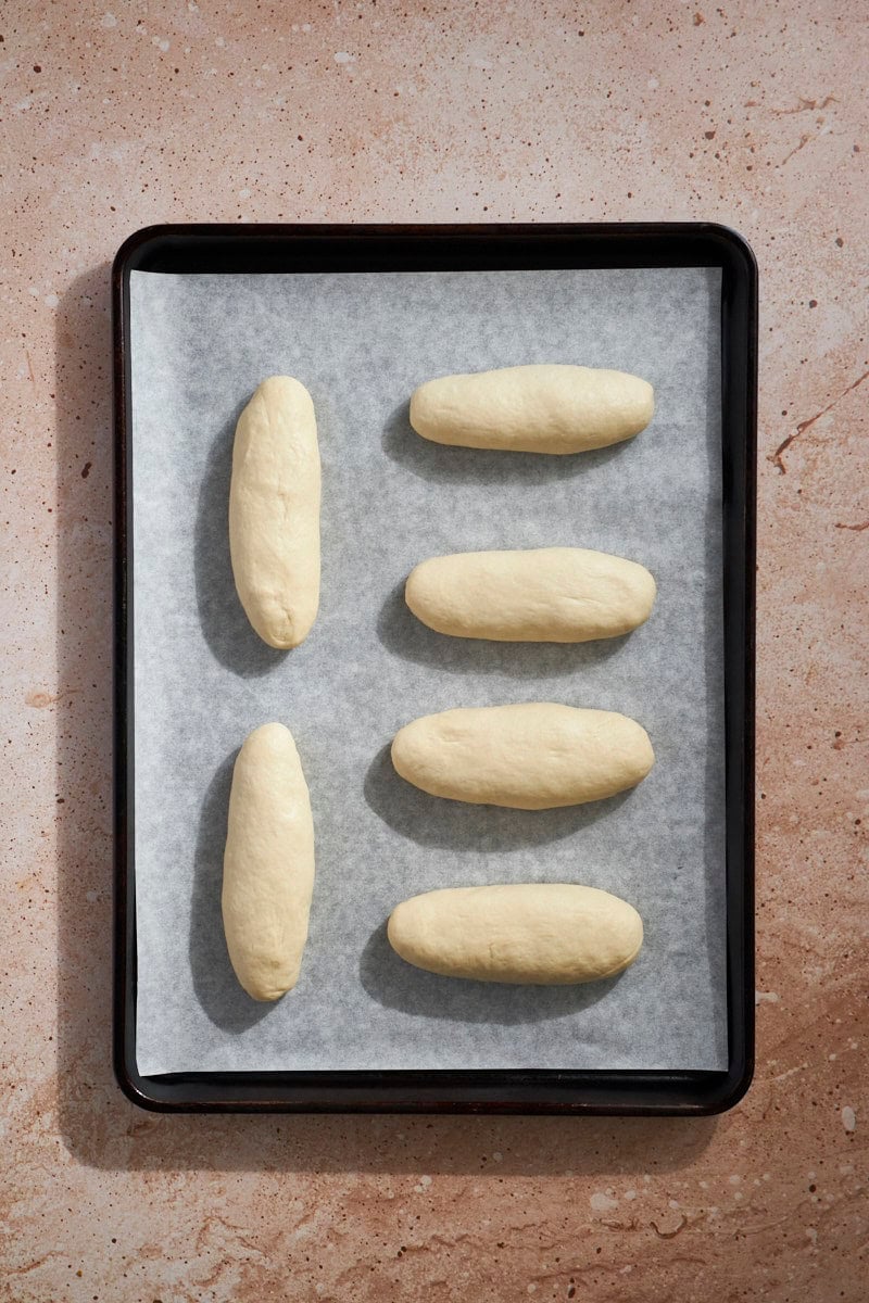 Buns shaped as hoagies on a baking sheet.