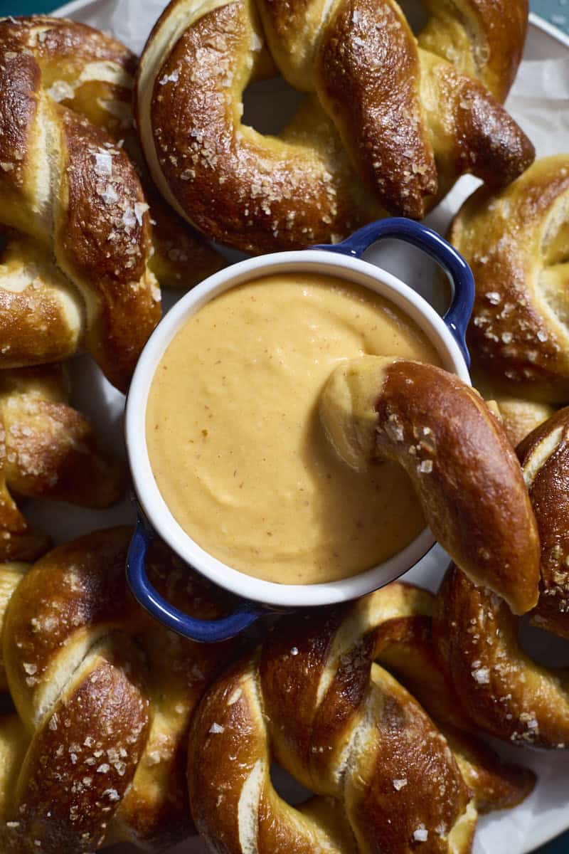 A closeup of a bowl of Beer Cheese on a platter of soft pretzels.