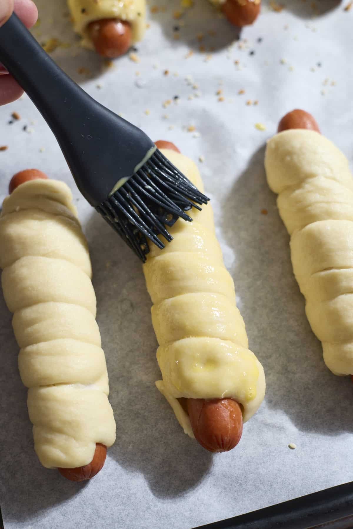 Egg wash being brushed onto a pretzel-wrapped hot dog.