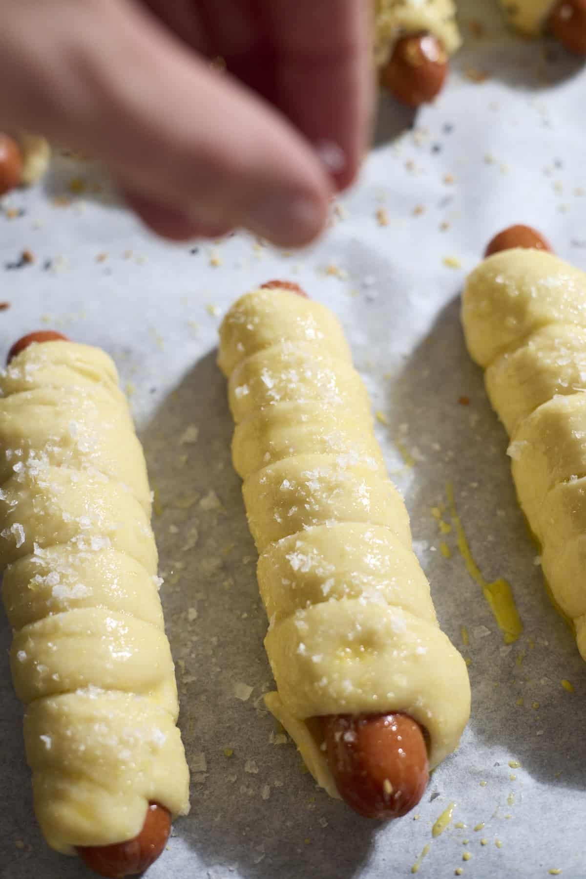 Salt being sprinkled on a pretzel-wrapped hot dog.